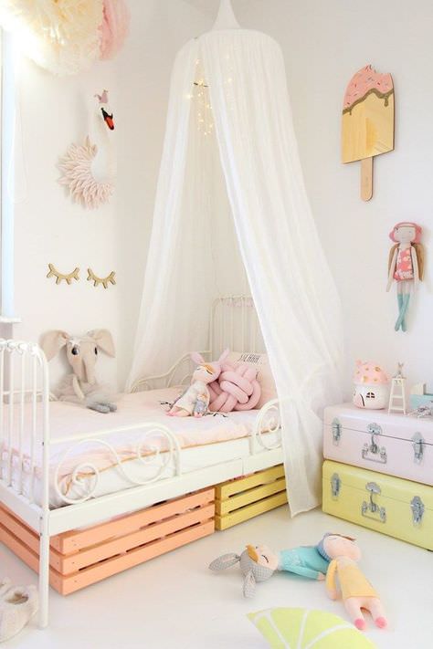 اتاق خواب کودک دختر با دیوارهای سفید که دارای تخت سفید، تور بالای تخت، میز کنار تختی چمدانی و عروسک های مختلف می باشد